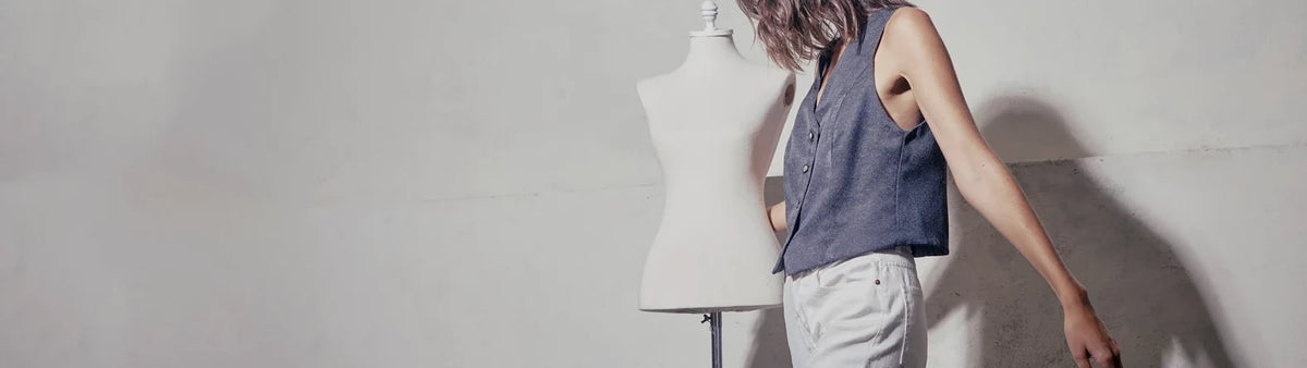 Woman in a blue vest and white trousers standing beside a dress form against a white wall